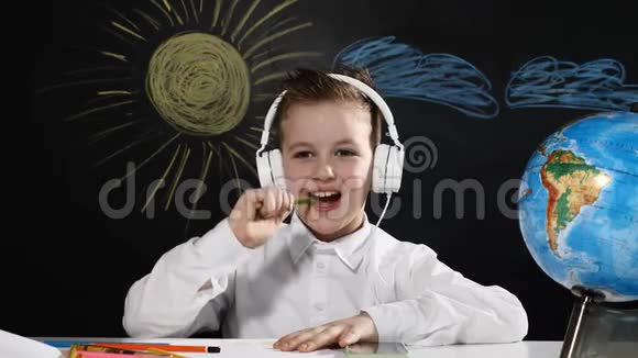 学校观念可爱的男孩坐在一张桌子上桌子上有地球仪后面有一块黑板带着耳机微笑的小学生视频的预览图