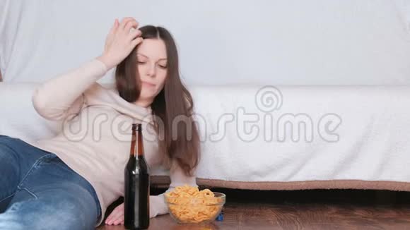 年轻漂亮的女人喝着啤酒在沙发附近的地板上吃着薯片念瓶子上的标签视频的预览图