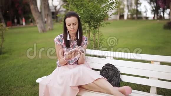 漂亮的女人坐在公园的长椅上用智能手机视频的预览图