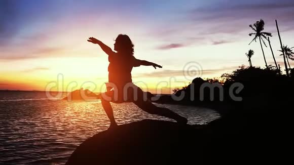 美丽的景色女人做瑜伽伸展在岩石和海景日落一个女孩在训练时摆姿势视频的预览图