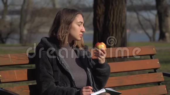 小女孩坐在公园的长凳上吃苹果在笔记本上写字视频的预览图