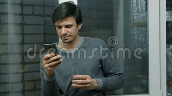男人正在用手机浏览网页站在阳台上的窗户附近喝着水视频的预览图