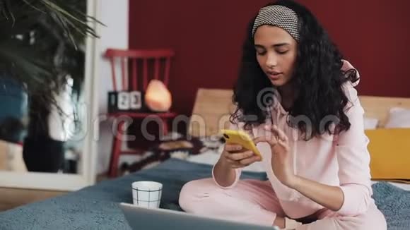 美丽的非裔美国女孩聊天和短信智能手机坐在床上在家里年轻女子视频的预览图