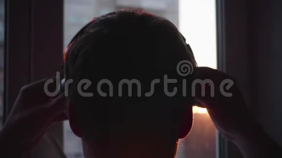 快关门一个年轻人站在窗户附近戴上耳机听音乐背景模糊日落视频的预览图