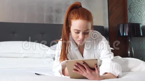 年轻的红发女人躺在酒店房间的床上用平板电脑看电影视频的预览图