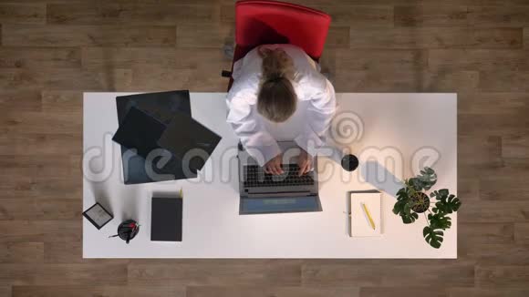 年轻的女医生坐在桌子上在笔记本电脑上打字喝着咖啡视频的预览图