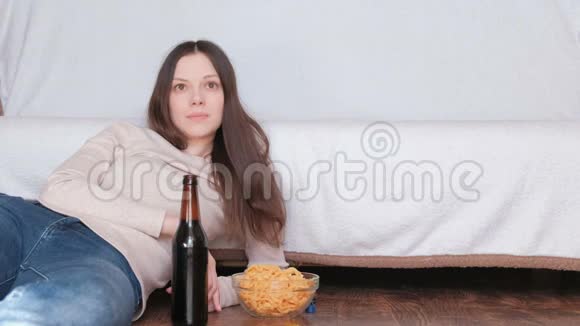 年轻的美女喝着啤酒吃着薯片躺在沙发附近的地板上看电视视频的预览图