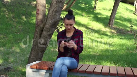 年轻漂亮的女孩穿着格子衬衫坐在公园的长凳上在网上聊天中积极改写视频的预览图
