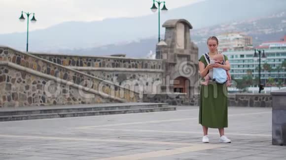 一个带着婴儿的年轻女人去欧洲旅行站在海边的广场上打电话视频的预览图