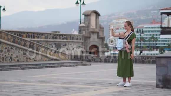 一个带着婴儿的年轻女人去欧洲旅行站在海边的广场上打电话视频的预览图