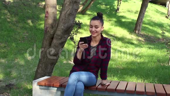 年轻漂亮的女孩穿着格子衬衫坐在公园的长凳上微笑着在网上聊天中积极地改写视频的预览图