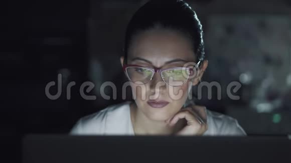 戴眼镜的女人在黑暗中在笔记本电脑上工作一个女孩晚上在电脑上消磨时间的肖像视频的预览图