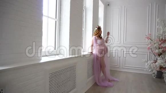 怀孕的女孩站在窗户附近穿着一件漂亮的粉红色连衣裙视频的预览图