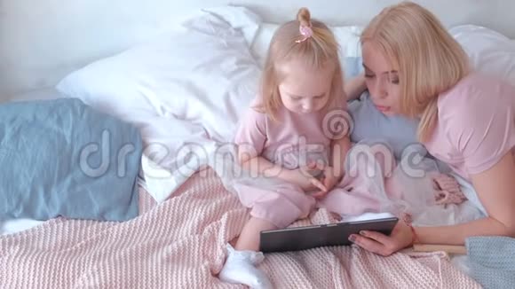 年轻迷人的金发女人在平板电脑上看书她的小可爱女儿穿着粉红色的连衣裙躺在床上视频的预览图