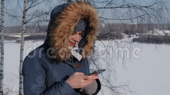 冬天公园里穿着蓝色羽绒服带毛衣罩的贴身女人在手机上写短信视频的预览图