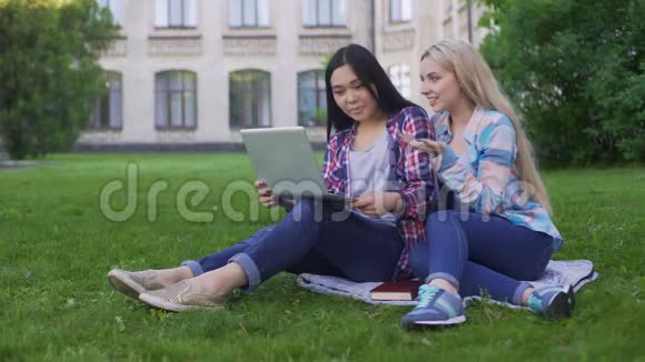 两个女学生坐在草地上在笔记本电脑上选择新衣服视频的预览图