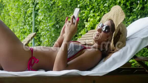 年轻漂亮的女士戴着太阳镜帽子和比基尼在游泳池旁边的躺椅上放松并使用移动工具视频的预览图