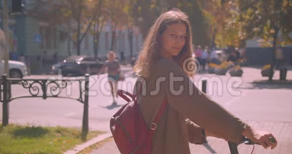 年轻可爱的白种人女性的特写镜头她站在户外的城市里用踢踏板车看着摄像机视频的预览图