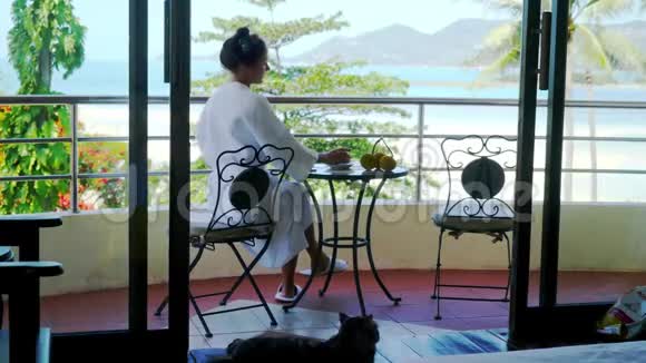 穿着浴袍的年轻女子坐在阳台上喝咖啡在令人惊叹的海景背景上使用手机视频的预览图