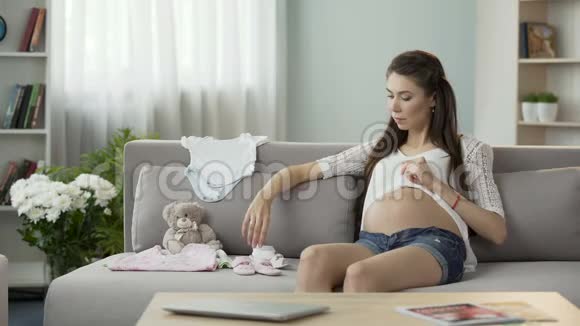 年轻孕妇看沙发上的儿童服装婴儿时尚视频的预览图