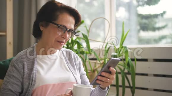 快乐的高级女人在家里坐在沙发上用智能手机视频的预览图