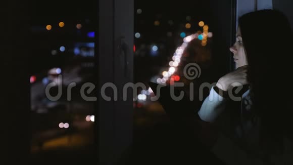 在黑暗的夜晚女人坐在窗台上看着街道平板电脑看书视频的预览图