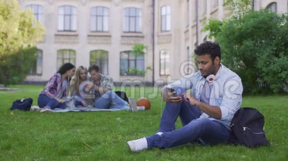 年轻的西班牙裔男学生坐在草地上在手机上滚动放松视频的预览图