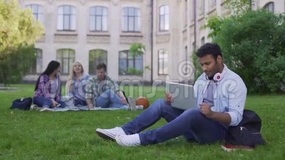 混合种族学生在笔记本电脑上上网聊天坐在大学附近的草地上视频的预览图