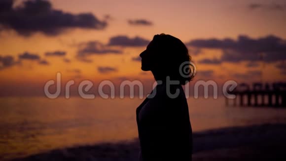 女人的剪影站在黄昏的海岸上在夕阳的背景下望着大海双手折叠视频的预览图