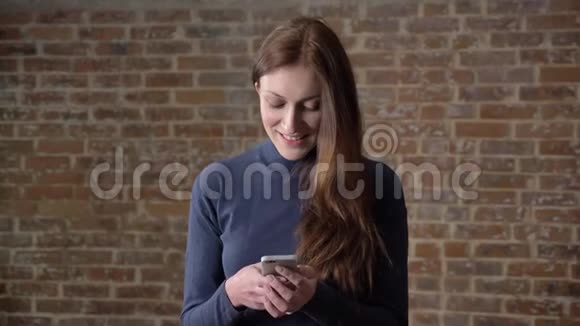 棕色头发的女孩正在智能手机上输入信息看着相机微笑交流理念砖背景视频的预览图