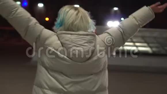 一位年轻的金发女郎穿着一件白色的时尚夹克晚上走在废弃的中央大街上视频的预览图