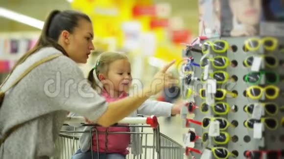快乐的女孩子与她美丽的母亲选择太阳镜在超市两个马尾辫的可爱女孩视频的预览图