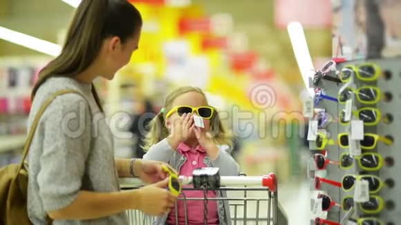 漂亮的小女孩正在超市里挑选太阳镜长着深色头发和黄色背包的女人视频的预览图
