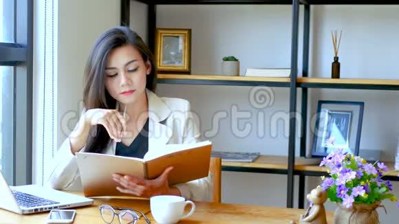 4K镜头美丽的亚洲商务女性坐在电脑笔记本电脑前阅读并翻翻笔记簿页面视频的预览图