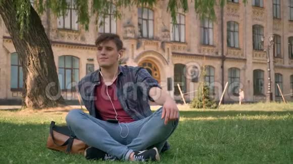 年轻的帅哥坐在公园的草坪上用耳机听音乐放松概念建立背景视频的预览图