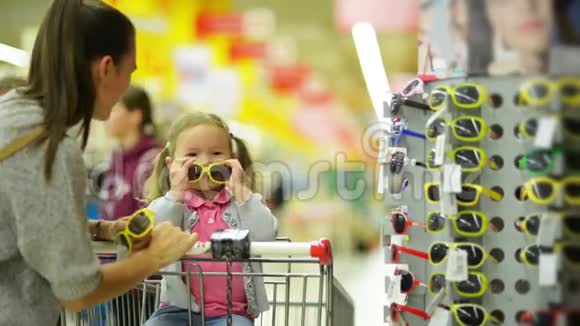 两条马尾辫的可爱丽蒂女孩坐在购物车里和她一起在超市挑选太阳镜视频的预览图