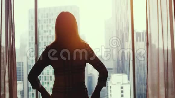 在美丽的夕阳下年轻的黑发女子站在大窗户旁望着城市建筑上的公寓视频的预览图