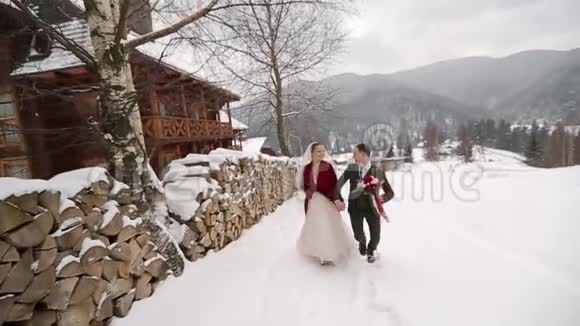 年轻的新婚夫妇跑着在滑雪胜地的村庄里玩着玩手里拿着木头小屋靠近原木冬季视频的预览图