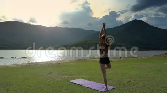 背面观景女孩站在瑜伽姿势垫与湖视频的预览图