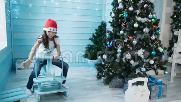 一个戴着红帽子的女孩坐在圣诞树附近儿童摇椅上视频的预览图