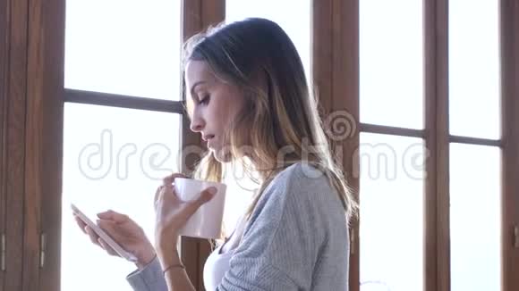 漂亮的年轻女人在家客厅的窗户附近喝咖啡时用手机视频的预览图