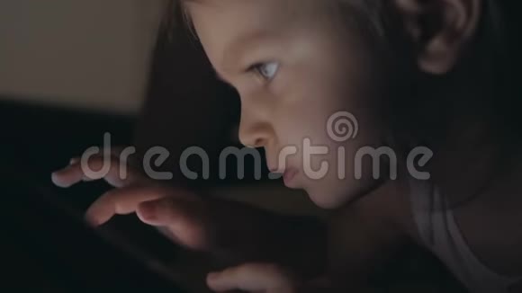 一个晚上躺在床上的小女孩在电子平板电脑屏幕上点击她的手指视频的预览图