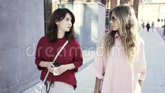 两个年轻的女人拿着咖啡杯在夏天的街道上散步和交谈视频的预览图