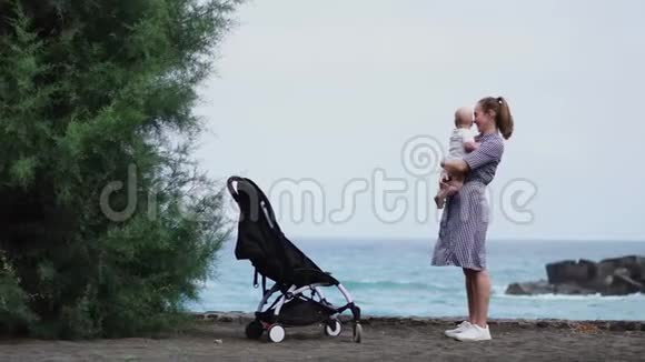 年轻母亲带着婴儿车坐在海滩上的后景视频的预览图