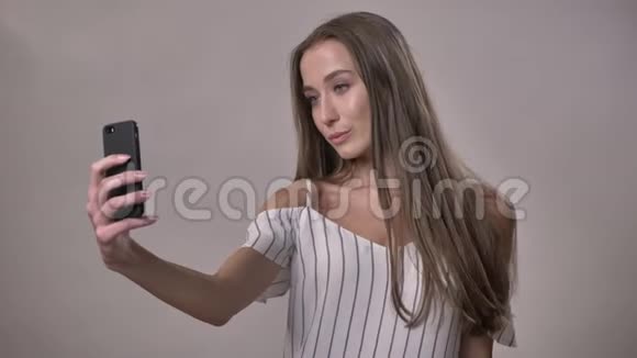 年轻迷人的害羞女孩正在智能手机上自拍交流理念灰色背景视频的预览图