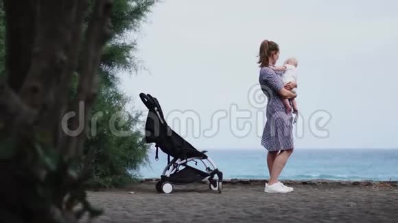 年轻母亲带着婴儿车坐在海滩上的后景视频的预览图