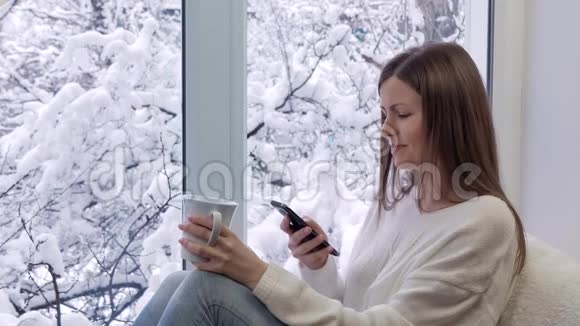 漂亮的女孩坐在窗台上喝茶用智能手机冬天在外面视频的预览图