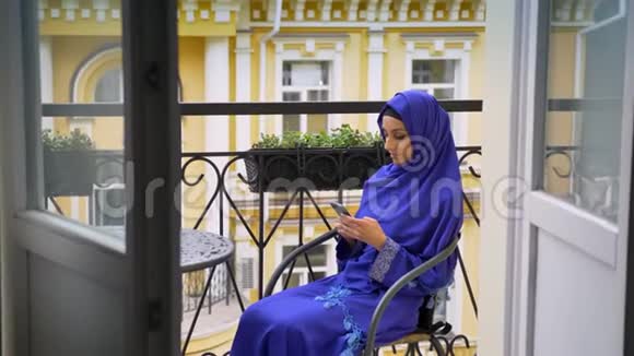 戴头巾的年轻穆斯林妇女坐在阳台上的椅子上打手机视频的预览图