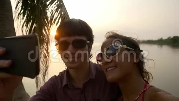 一对年轻快乐的夫妇戴着太阳镜在浪漫的节日里在热带海滩上坐在手掌上自拍视频的预览图