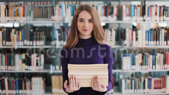 微笑的女学生拿着书站在图书馆的书架前视频的预览图